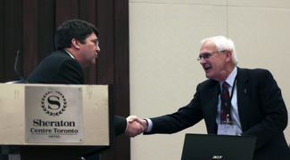 Two men, both wearing sport jackets, lean toward each other to shake hands. They are partly obscured by the top of an open laptop on a table and by a large podium bearing the words "Sheraton Centre Toronto"