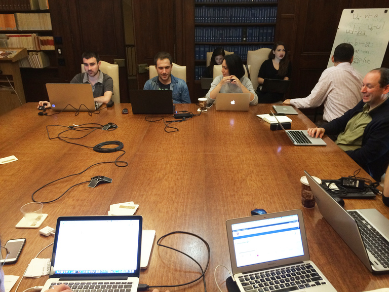 A group of people site, each with an open laptop, around a large square wooden table in a wood-paneled room with bookshelves. Some seem to be working on their computers, others are talking to each other. Two people in the corner are conferring over text written on a whiteboard.