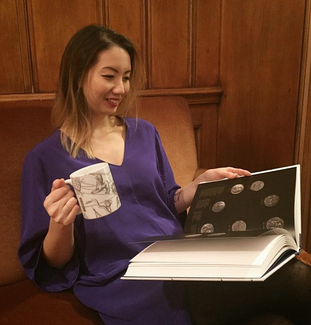 A smiling woman sits on a couch or bench in a wood-paneled room. In one hand, she holds a large, white mug with figures and lines on it in dark gray ink. In her lap is a large book that she holds open with her other hand. A entire page can be seen: it features seven round objects arranged in a circular pattern on a dark background.