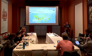 A group of people sit in a large room at a U-shaped arrangement of tables. A man in a sport coat stands at a podium in the front of the room and gestures toward a large projection screen on which is depicted a map of Eurasia, with irregular blotches of color in various areas including much of southern and central Europe, the Middle East and portions of Central Asia and China.