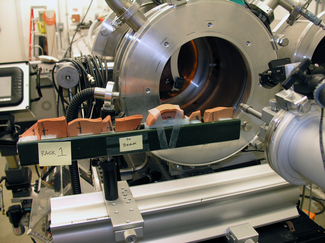 In this photograph, pieces of terracotta-colored ceramic are lined up in a metal tray in front of a complex metal laboratory device consisting of a large metal cylinder behind and a smaller cylindrical device with attached components in the front.