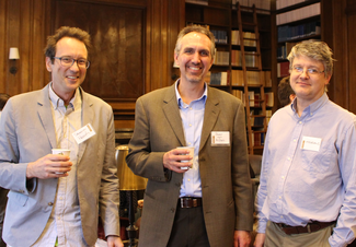 Three people with name tags smile for the camera. Two are holding coffee cups. They are standing in a wood-paneled room with book shelves.