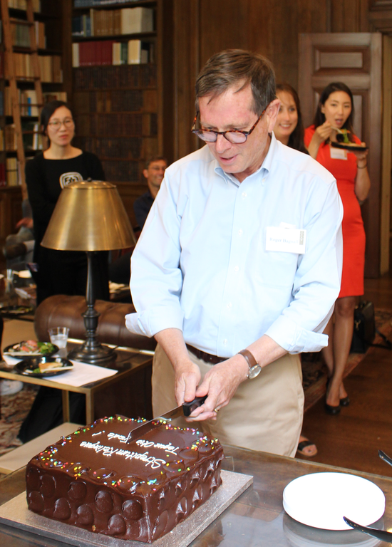 A man smiles and cuts a large, chocolate cake while others stand and sit behind him, smiling. They are in a wood-paneled room with bookshelves.