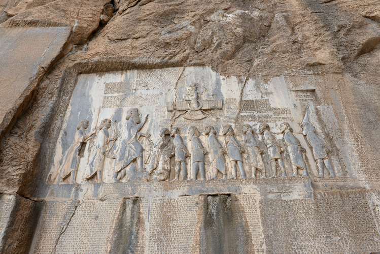 Photo of a rock-cut inscription depicting multiple figures in line with their hands bound behind their backs facing a larger individual who raises a hand. Cuneiform writing appear around and on the figures.