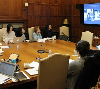 Several people with laptops and notebooks sit at a large square wooden table in a wood-paneled room. One one wall, a large video screen shows a series of indistinct black-and-white photographs.