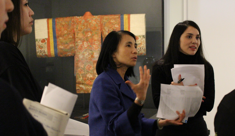 Two women holding papers stand in front of a large panel displaying an article of colorful Asian dress, speaking to other people gathered nearby.