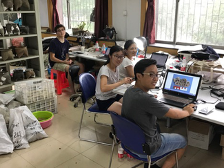 Four people sit at a series of worktables with laptops in a large room with metal shelves and windows. On the shelves are a number of ceramic objects. Plastic crates and polymer bags are lined up on the floor.