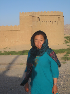 A woman poses in front of what appears to be a contemporaneous or restored mud-brick fortification wall and tower.