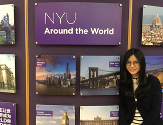 A woman poses in front of a display that carries a large title "NYU Around the World". The rest of the display is dominated by photographs of cities around the world.