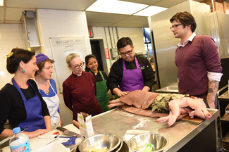 Six people, some wearing aprons, gather around a large work surface in a kitchen. On the work surface is a whole pig. It has been gutted and is wrapped in green leaves. One individual is covering part of it with a brown, doughy material that has been rolled out flat.