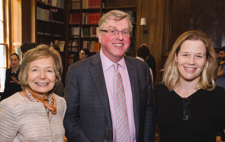 Three people pose for photograph in a wood-paneled room lined with bookshelves. Other people can be seen behind them, conversing and part of a large window can also be seen.