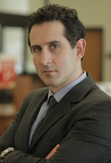 A man in a suit and tie stands at an angle to, but looking into, the camera with his arms folded.