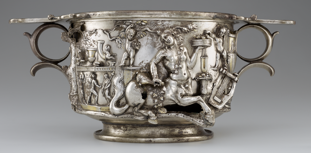 An elaborately decorated silver cup, shaped like a cylinder with two round handles opposite each other just below the lip. The cylindrical cup sits on a slightly smaller base. A female centar, puti, other small figurrees, and musical instruments can be seen in the decoration.