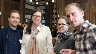 Four individuals pose for the photograph in a room with wood paneling, book shelves, and elaborate pendant light fixtures. They are wearing name tags, but the text on them cannot be read.