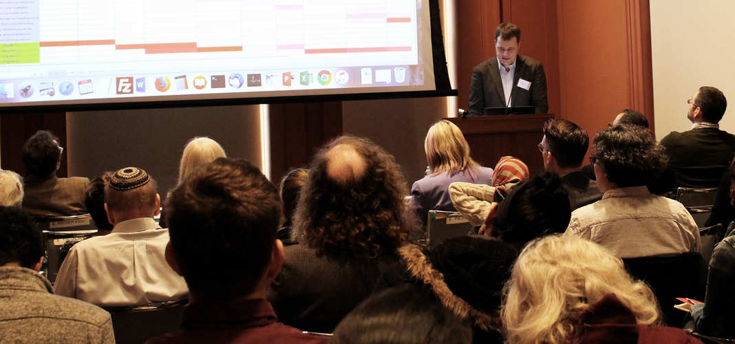 A photograph taken from the rear of a full lecture hall. Several seated audience members can be seen from behind. Beyond them, a man in a sport jacket stands at a podium facing the audience. Next to him is a large projection screen showing textual data in tabular form.