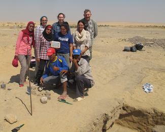 Several people pose next to a trench on an excavation site in a desert area.