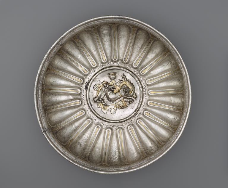 Picture of a shallow, decorative bowl made of silver and gold. In the center is a medallion depicting a woman sleeping on a lion skin, surrounded by a club and other objects.