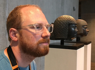 Photograph of Christian Casey posing in a museum next to two egyptianizing sculptural heads.