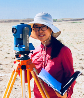 A woman stands behind an optical survey device on a tripod in a desert landscape. She wears a sun hat, glasses, and holds an open notebook in one hand.