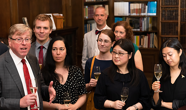 Several people hold wine glasses and stand together in a room with bookshelves.