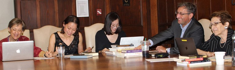 Four women and a man are seated at a large table in a wood paneled room. Several are taking notes. Laptop computers and books are in use on the table-top.