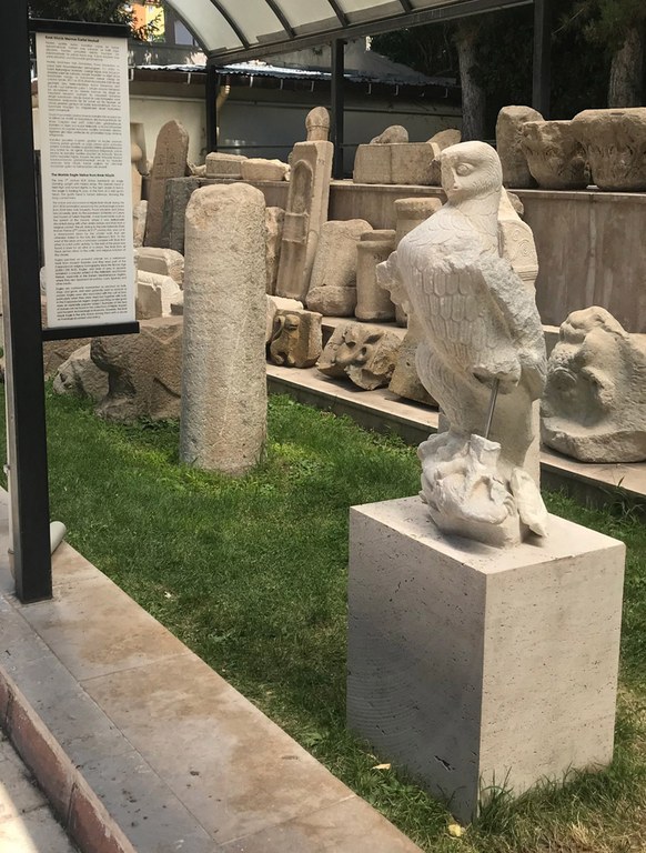 A stone statue of an eagle stands on a cubic stone base. Numerous other stone artefacts appear in the background.