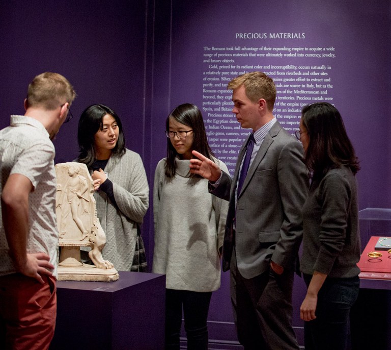 Five people stand talking in an ISAW gallery next to a carved stone object. Text on the wall behind them reads: "Precious Materials."