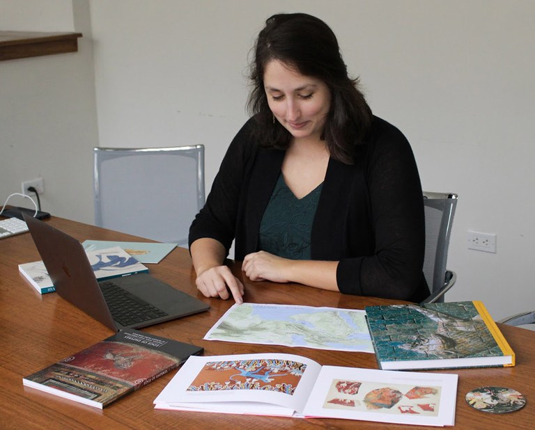 A woman sits at a desk, looking down at a map. A laptop and a book are also open on the table top, and a number of other books are arrayed around her.