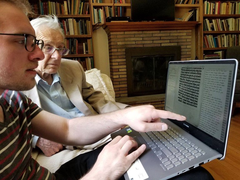 Two men sit looking at a laptop. One man points at text on the screen. The wall behind them is covered in book cases full of books, apart from a fireplace in the center. 