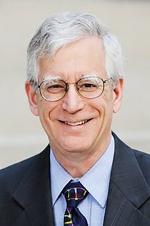 A man wearing glasses and a suit and tie smiles into the camera.