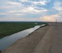 Photograph of a water-filled ditch stretching into the distance. On the left is a green, apparently cultivated field. On the right: packed dirt, possibly a roadway.