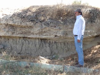A man stands in profile in front of an excavated section of earth almost as tall as he is. Several stratigraphic layers are visible.