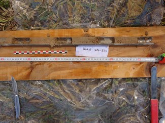 On a wooden board, a core-sampling tube is laid out along with tools, a ruler, and a scale bar. Sections of dirt are visible in the tube.