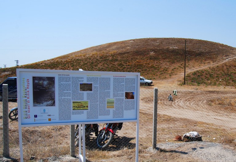Photograph of mound of Niğde-Kınık Höyük with welcome information panel in the foreground and blue sky in background.