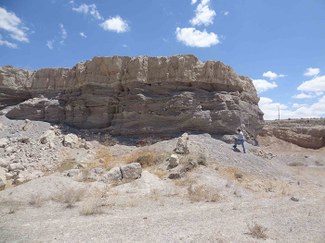 In a desertified landscape with blue sky beyond, a man stands in front of a large vertical rock outcropping four or five times as tall as he is.